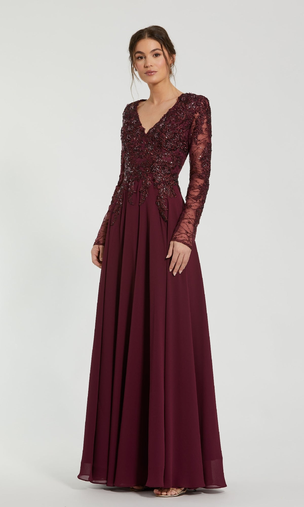 A woman wears the Mac Duggal 20388 long sleeve special occasion dress, a floor-length burgundy gown with lace and sequin details, standing against a plain light background.